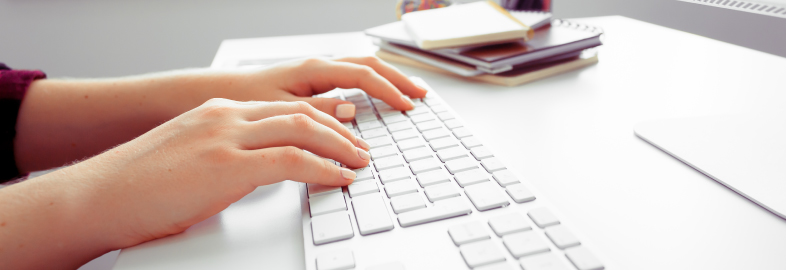 Manos escribiendo en teclado blanco con cuadernos apilados en escritorio, actividad digital de trabajo o gestión.